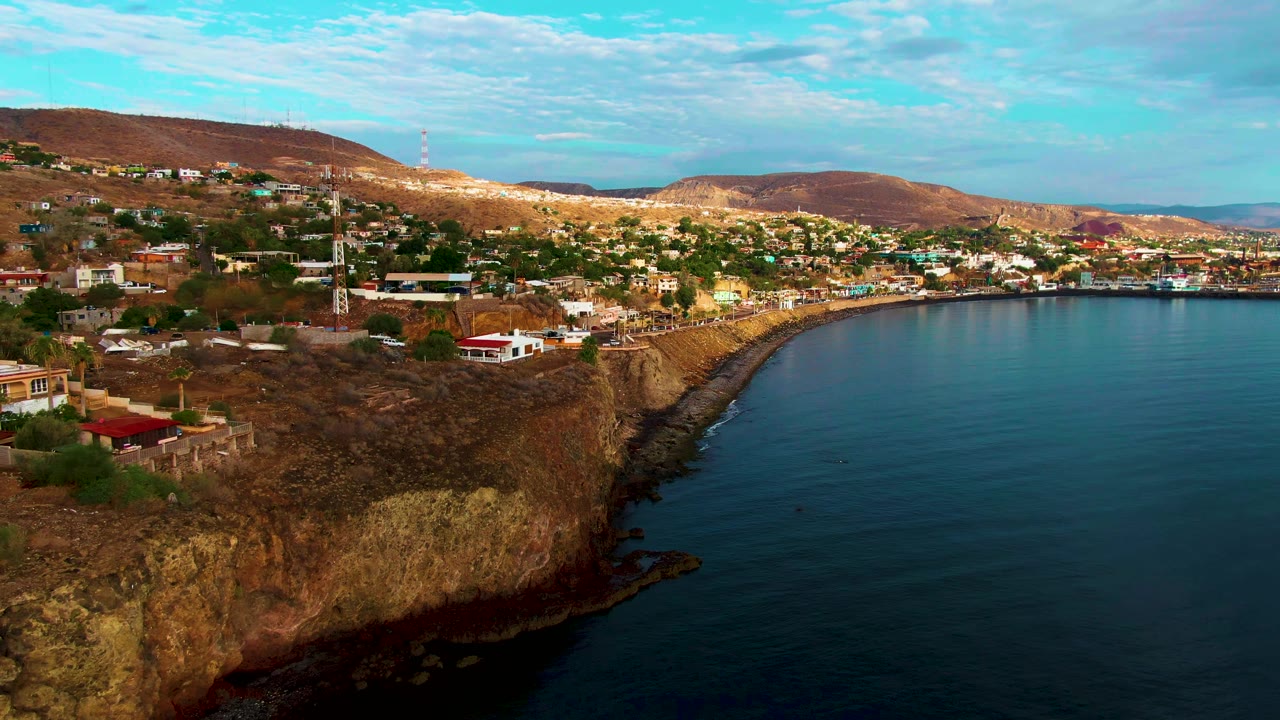 Aerial view of Santa Rosalia, Baja California Sur, with cliffside houses above dark blue ocean water, dry hills in the background, scattered clouds, and no visible people.