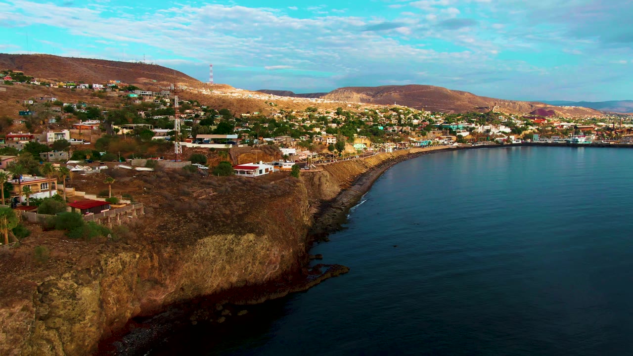 Santa Rosalia coastal buildings and a tall tower sit above a rugged cliff line with the bay to the right, dry hills beyond, and no visible people.
