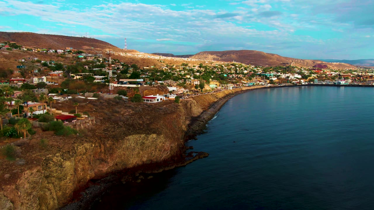 The aerial view shows Santa Rosalia houses following the cliff curve around calm blue water, with warm light on the hills and no visible people.