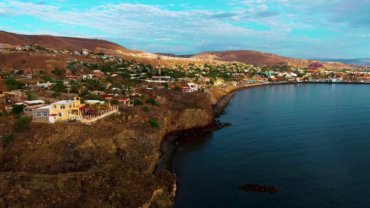 Santa Rosalia cliffside homes and dry hills overlook a calm blue bay at the opening moment, with no visible people.