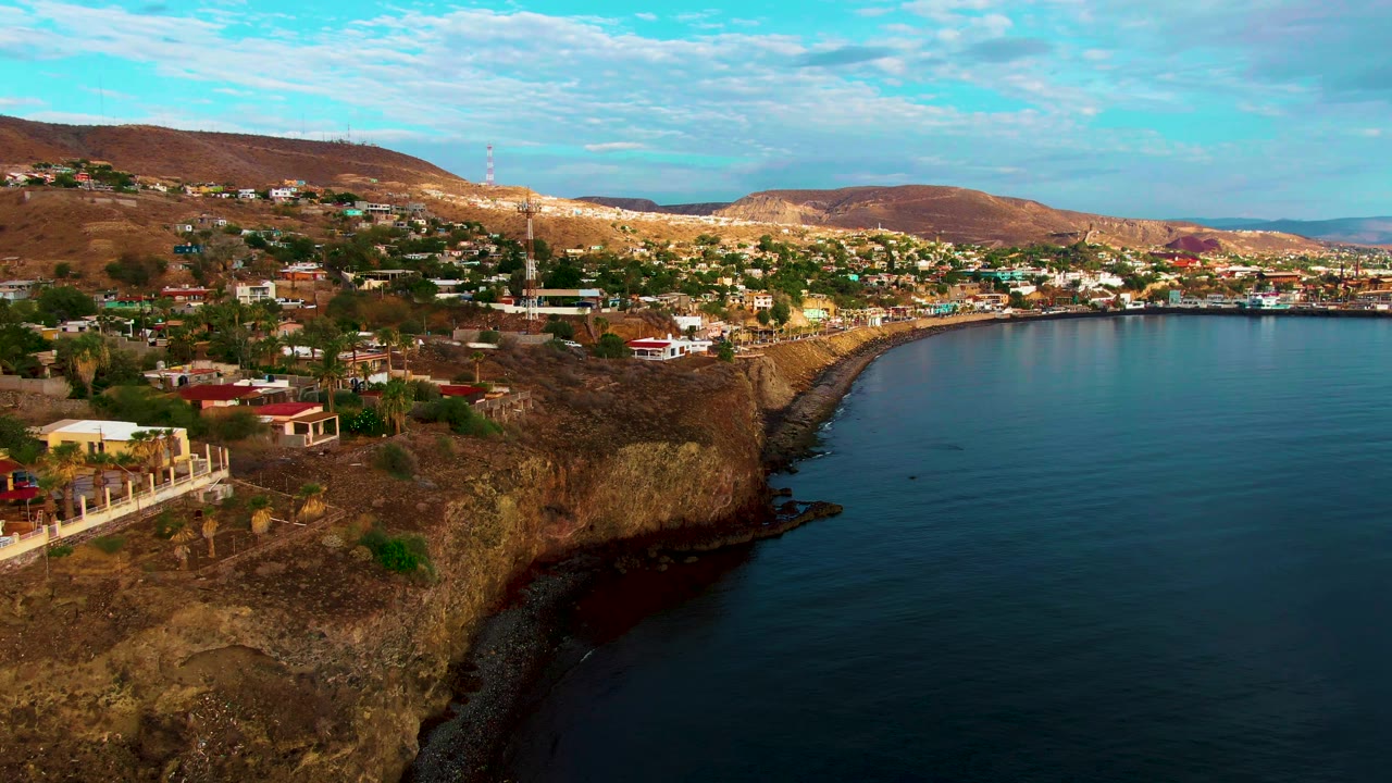 Opening aerial frame over Santa Rosalia, Baja California Sur, showing hillside buildings on tan rocky cliffs above blue ocean water with no visible people.