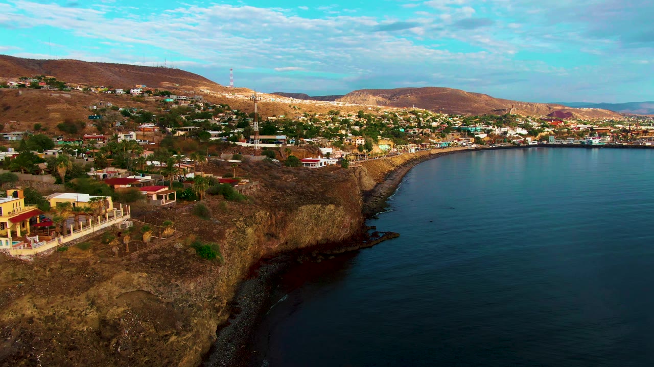Coastal town buildings spread across dry hills above a rocky ocean edge.