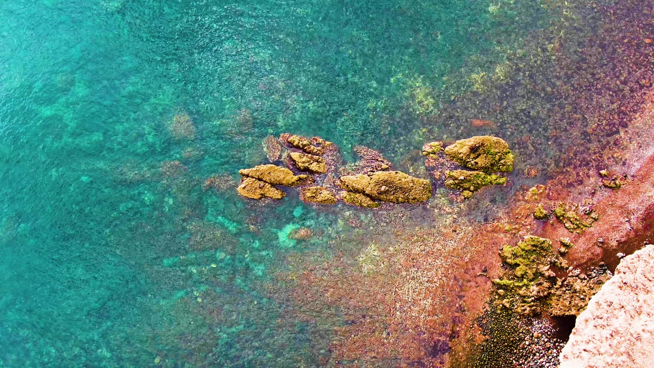 Overhead view of shallow turquoise water lapping around mossy rocks beside a red-brown Santa Rosalia shoreline, with no visible people.