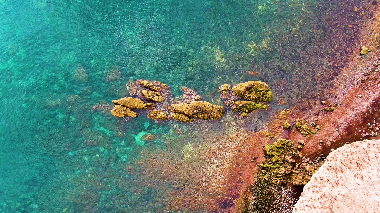 An overhead frame shows shallow turquoise water washing around a compact rock cluster near the red-brown Santa Rosalia shore, with no visible people.