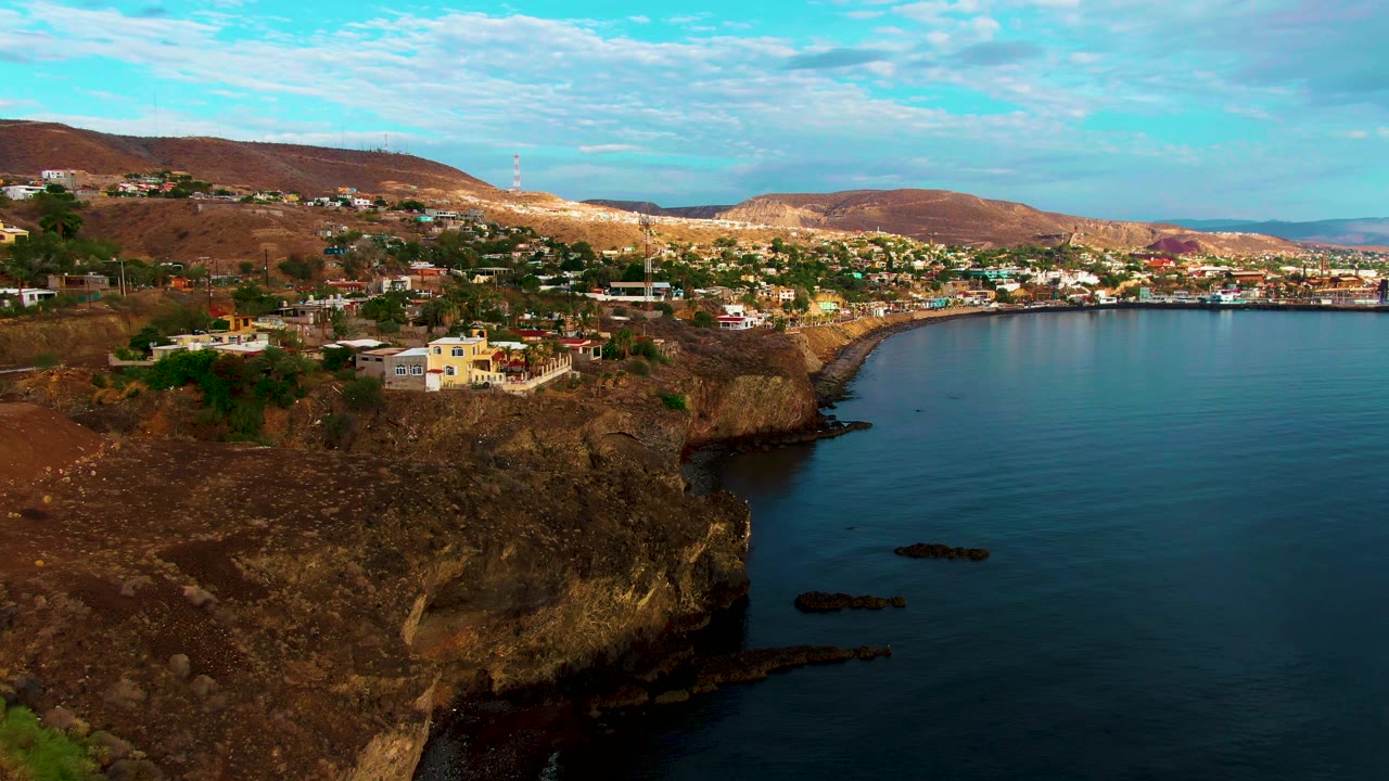 Wide aerial frame over Santa Rosalia, Baja California Sur, showing brown cliffs with scattered buildings, calm blue ocean to the right, town along the bay, low hills, scattered clouds, and no visible people.