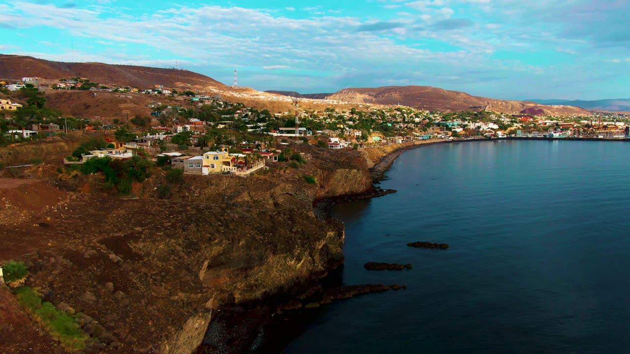 Aerial view in Santa Rosalia, Baja California Sur, with more of the cliff face centered, small buildings on the slope, calm ocean on the right, a town band across the bay, and no visible people.
