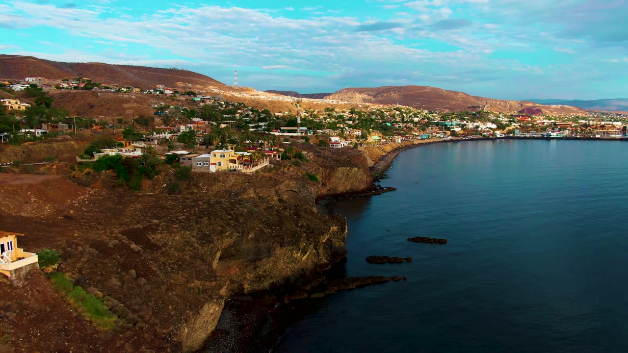 Aerial frame over Santa Rosalia with the rocky headland pushed farther left, a small pale building near the lower edge, dark rocks offshore, calm blue sea, distant town, and no visible people.