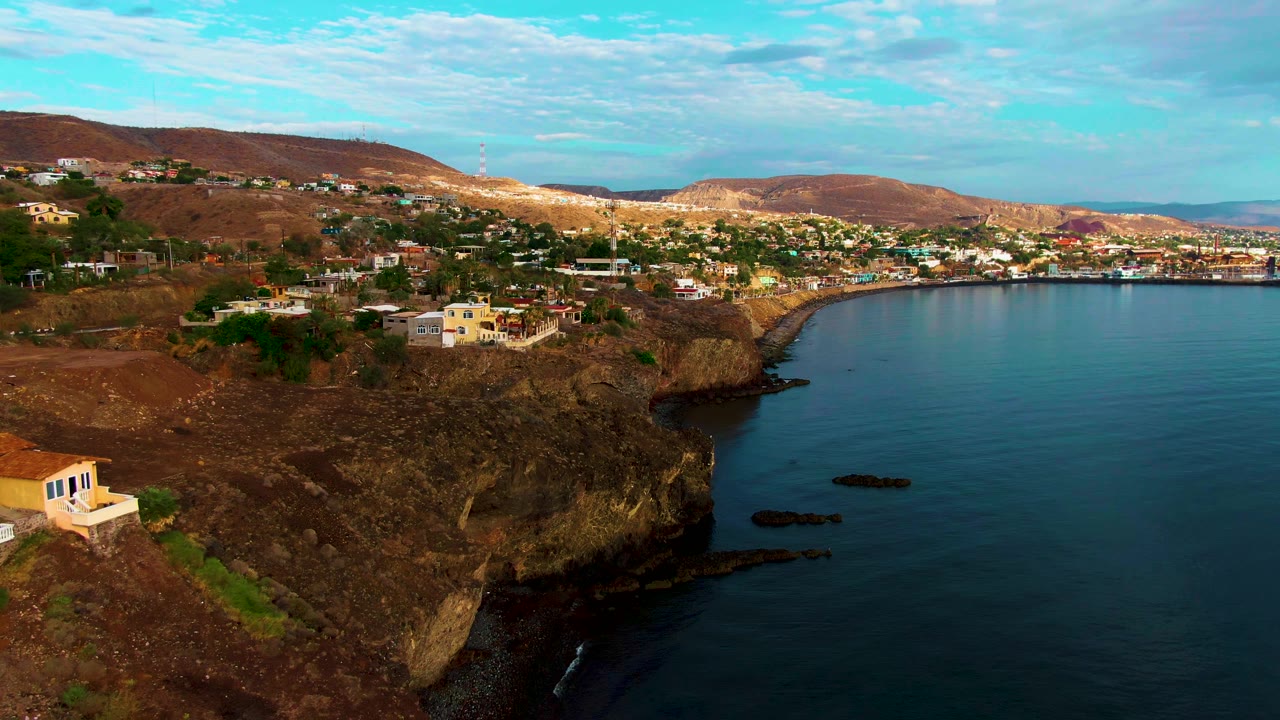 Wide coastline aerial with cliff, bay water, hillside buildings, and desert hills