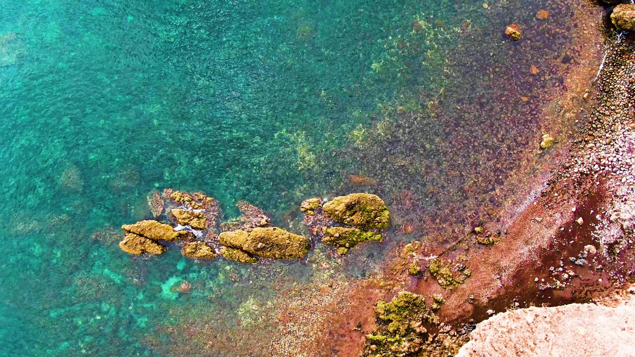 Clear turquoise water surrounds a low mossy rock cluster beside reddish wet shoreline in Santa Rosalia, Baja California Sur, with no visible people.