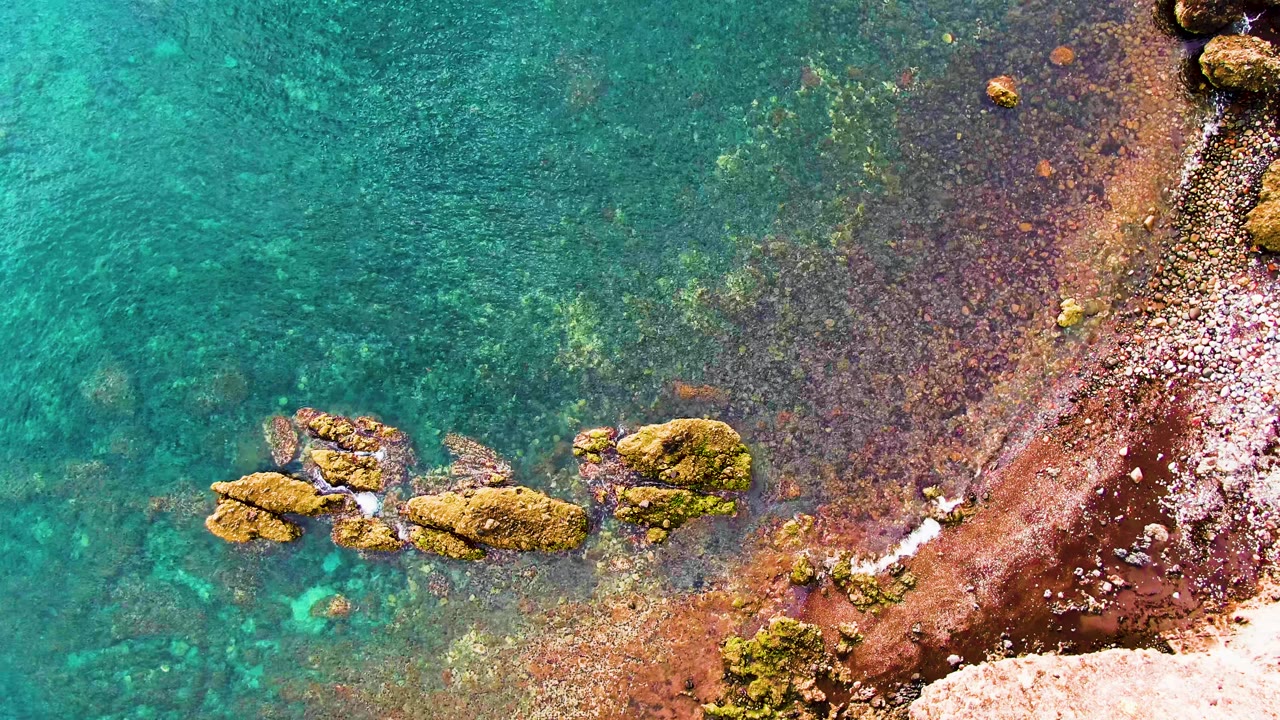An overhead coastal frame shows mossy rocks in shallow turquoise water with a curved reddish shore and pale pebbles in Santa Rosalia, Baja California Sur, with no visible people.