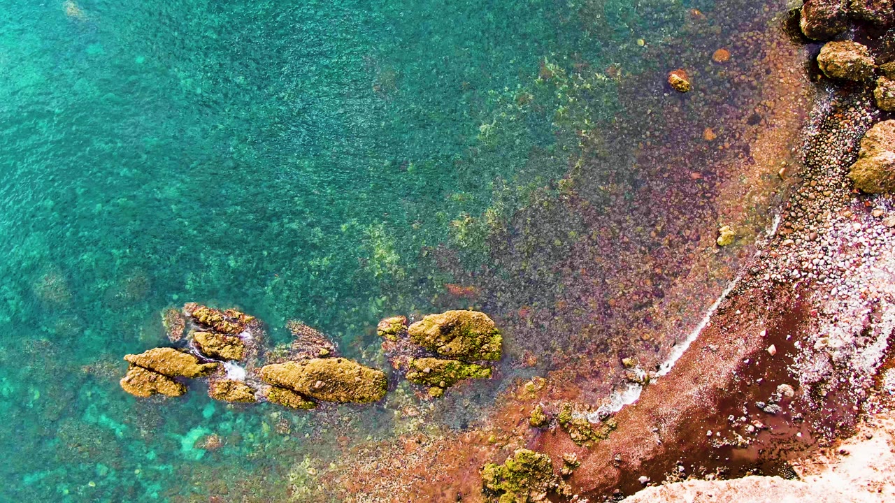 Turquoise water covers the left of the frame while gentle surf traces the reddish rocky shore on the right in Santa Rosalia, Baja California Sur, with no visible people.