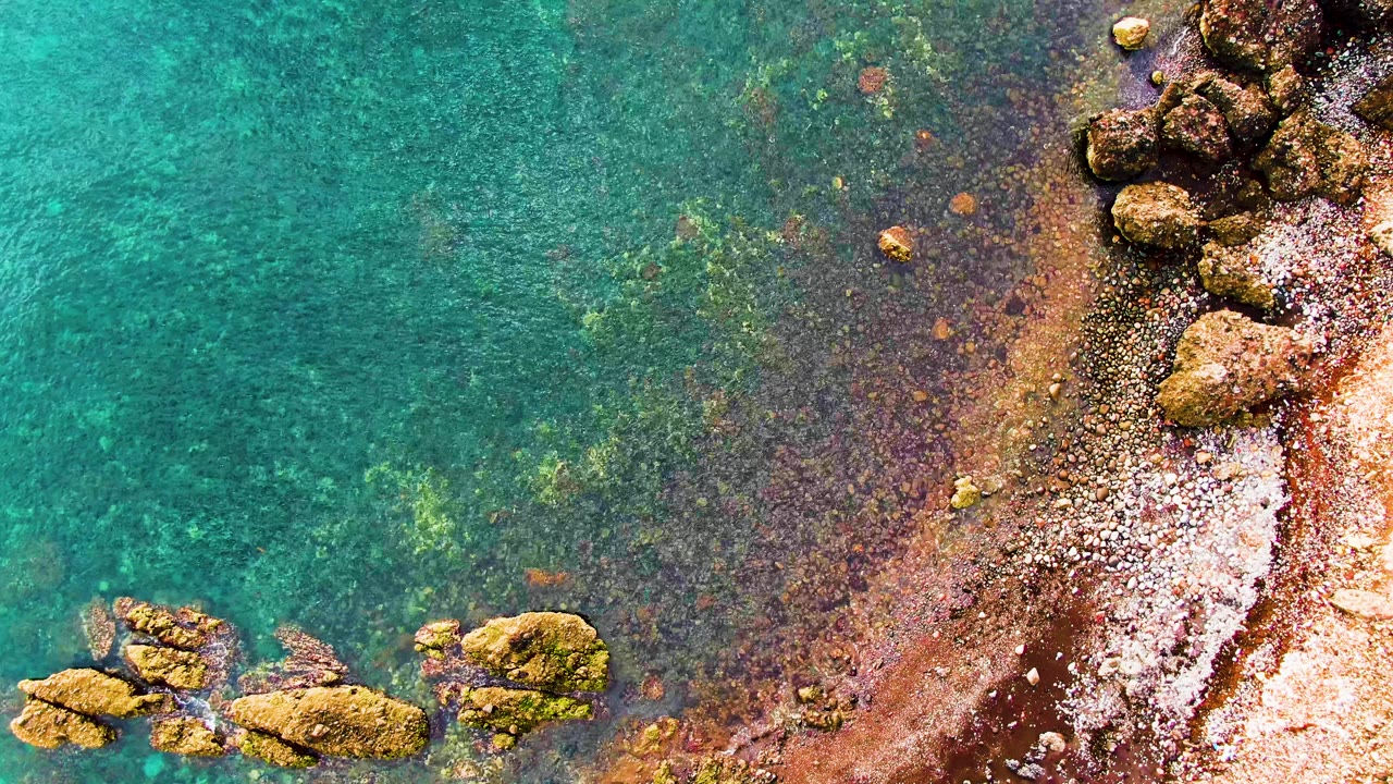 Overhead view of clear turquoise water meeting a rocky sandy shoreline in Santa Rosalia, Baja California Sur, with no visible people.