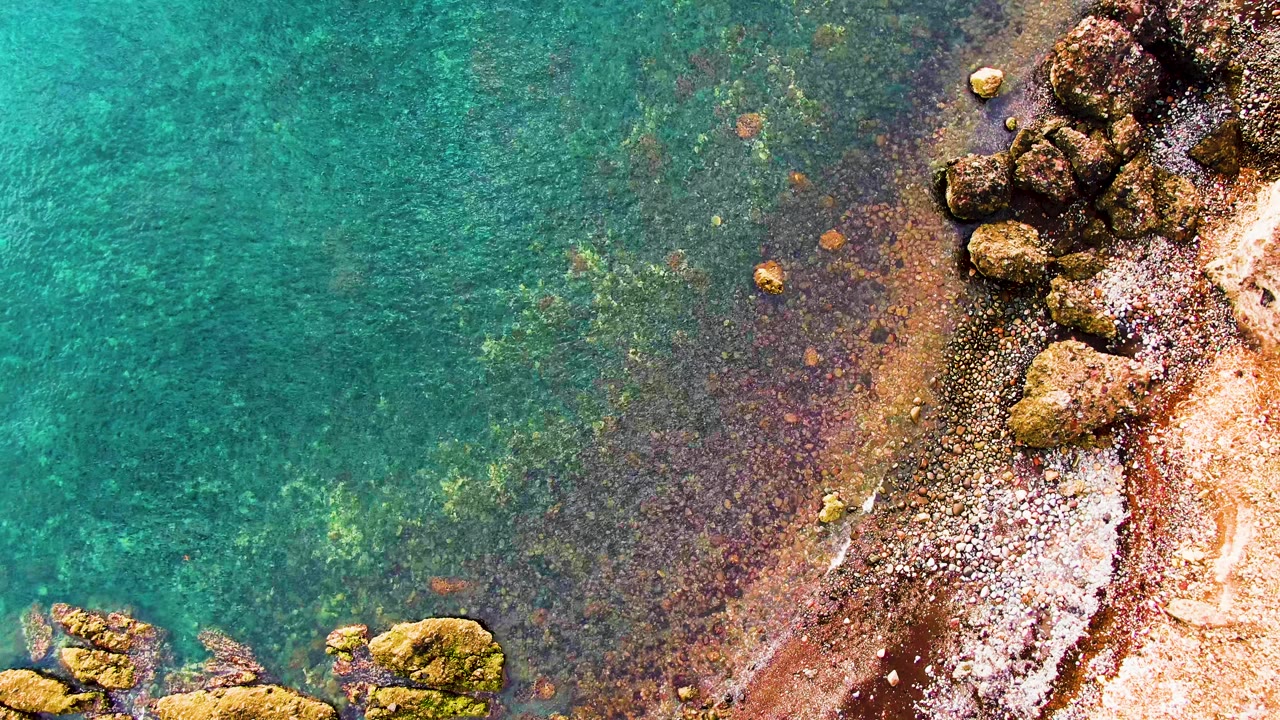 Aerial frame of tan rock outcrops, reddish sand, and clear water along the Santa Rosalia coast, with no visible people.