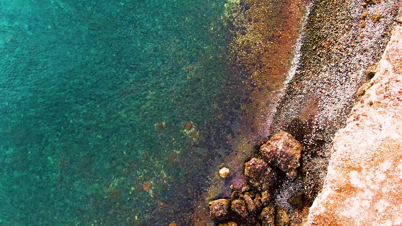 Top-down view of turquoise water meeting a rocky Santa Rosalia shoreline, with pebbles, pale rock, and no visible people.