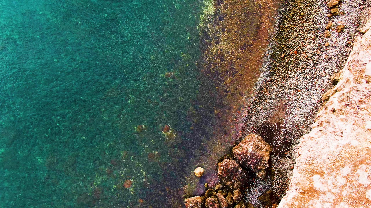 Clear turquoise sea laps against pebble bands and sunlit rocks along the Santa Rosalia coast, with no visible people.