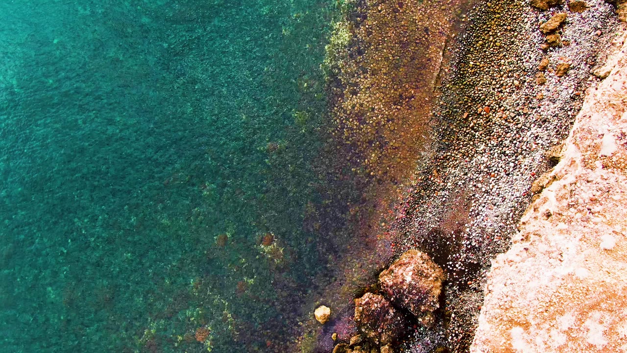 Aerial frame over teal water and a narrow rocky beach in Santa Rosalia, with scattered foam patches and no visible people.