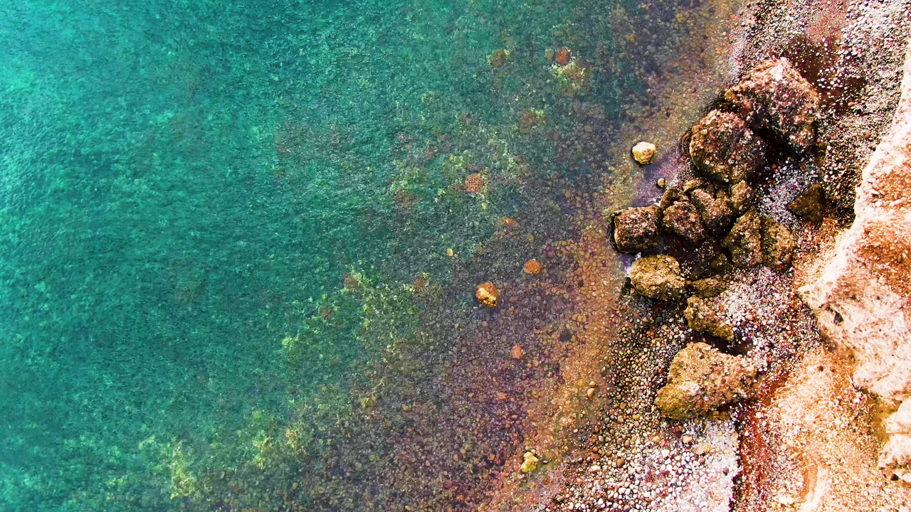 Overhead view in Santa Rosalia, Baja California Sur, of clear turquoise water meeting a rocky shore along the right edge, with submerged rocks visible and no visible people.