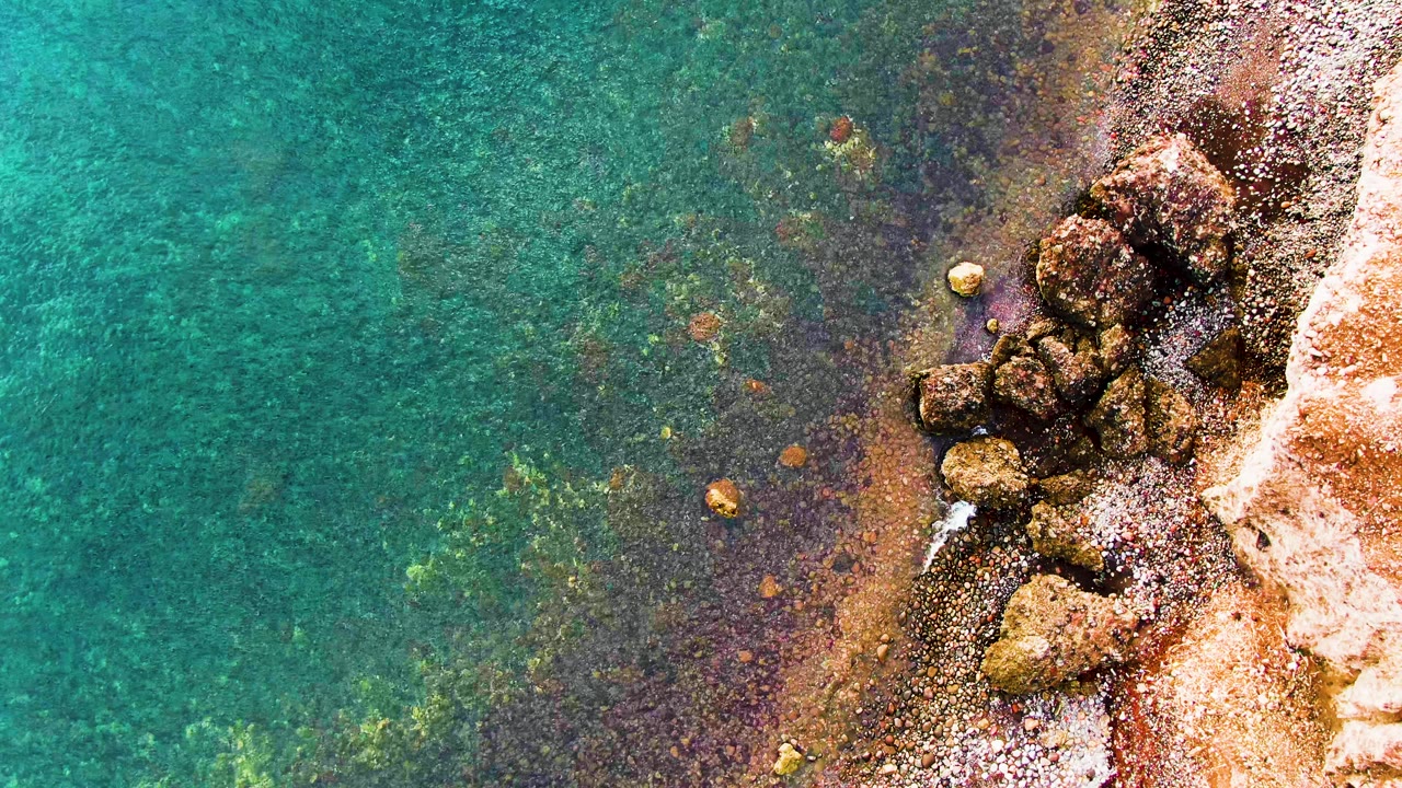 Overhead Santa Rosalia coastal frame showing transparent green-blue water over seabed rocks, with a clustered rock outcrop and pale surf line on the right, and no visible people.
