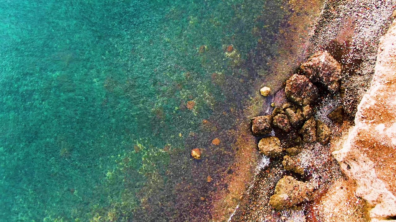 Top-down view of Santa Rosalia shoreline where turquoise water darkens over submerged rock patches and small surf touches the pale rocky edge, with no visible people.