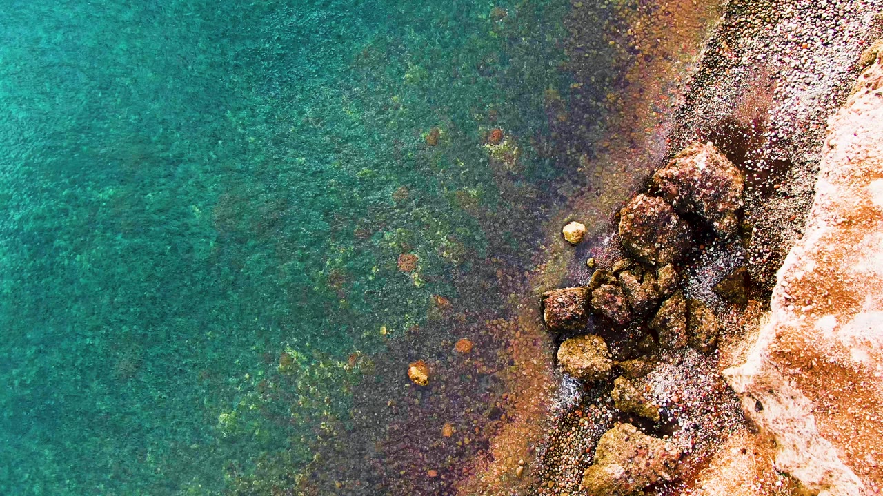 Overhead Santa Rosalia view with green-blue water covering the left side and a tight cluster of shoreline boulders on the right where small waves lap, with no visible people.