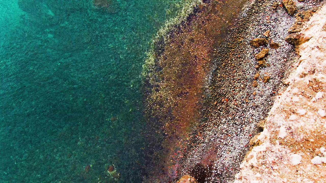 Top-down aerial view of turquoise water meeting a diagonal rocky shore in Santa Rosalia, Baja California Sur, with pale land at right and no visible people.