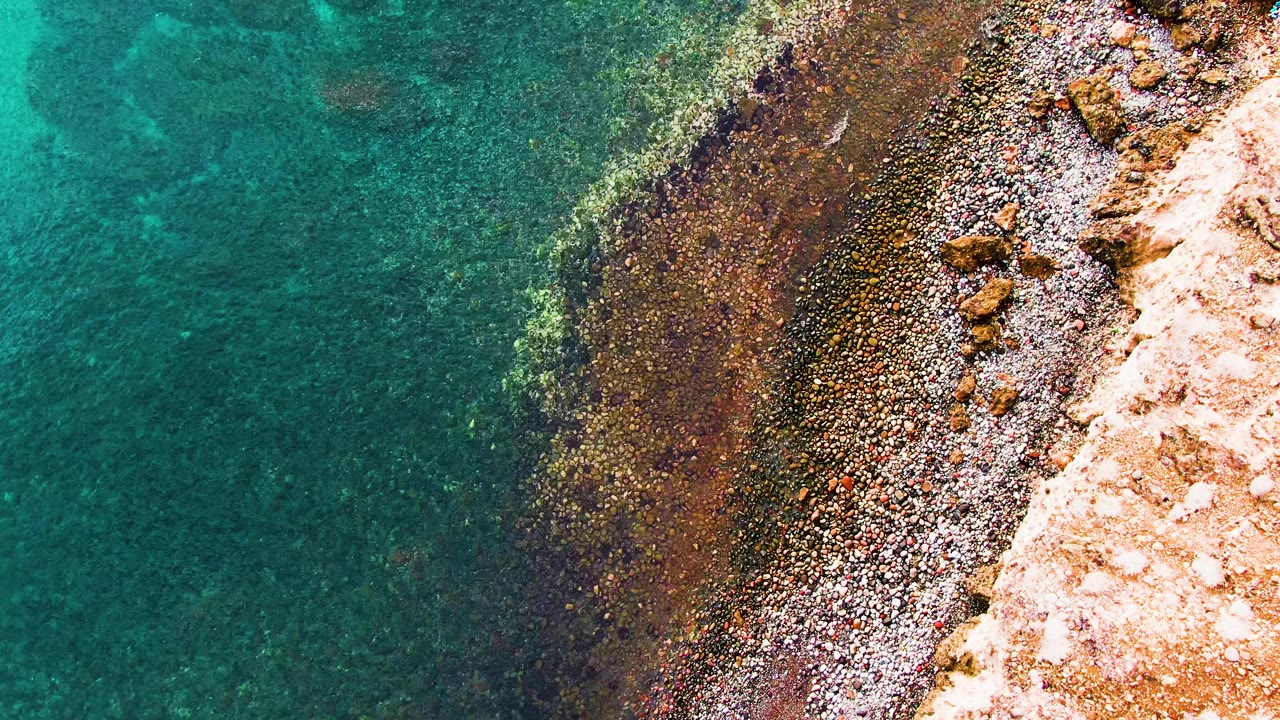Overhead view of Santa Rosalia coastline with turquoise water, a dark shallow-water rock shelf, scattered pebbles, pale shore rock, and no visible people.