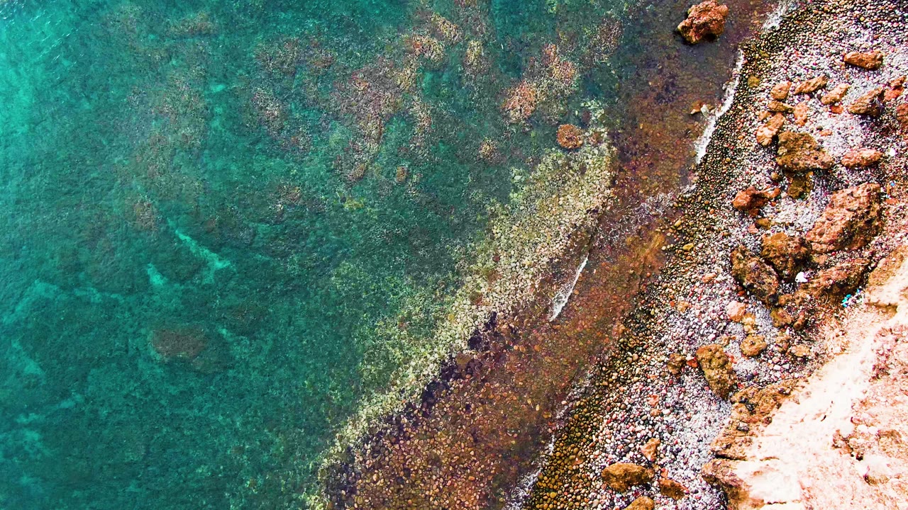 Aerial view over Santa Rosalia shallow water where a narrow white wave line traces the stony coast and reddish rocks sit near the right edge, with no visible people.
