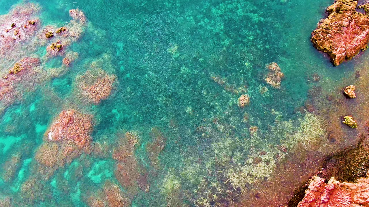 A top-down Santa Rosalia frame shows turquoise shallow water, mottled seabed color, reddish rock formations, and no visible people.