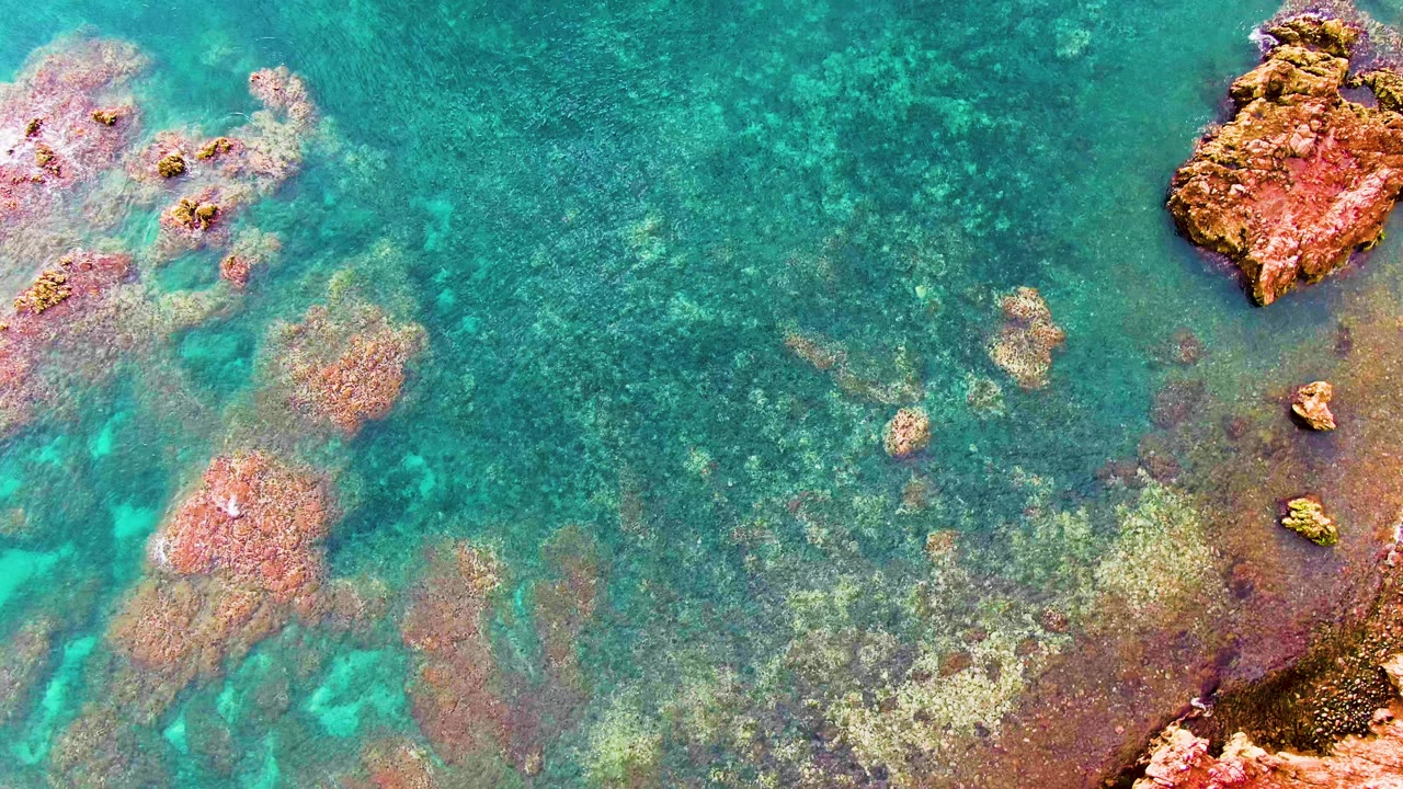 Clear Santa Rosalia water reveals reddish submerged rocks and pale seabed patches, with larger rock edges visible and no visible people.