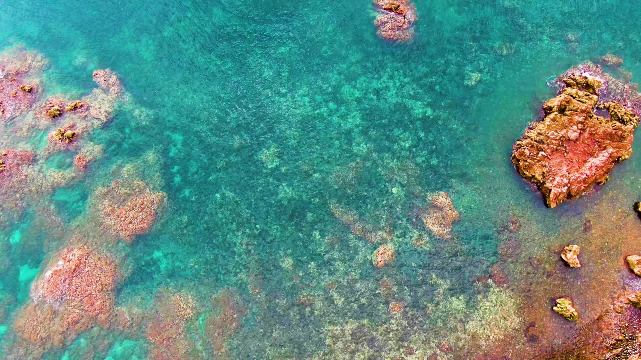 Turquoise shallow water with reddish rocks and submerged seabed texture.