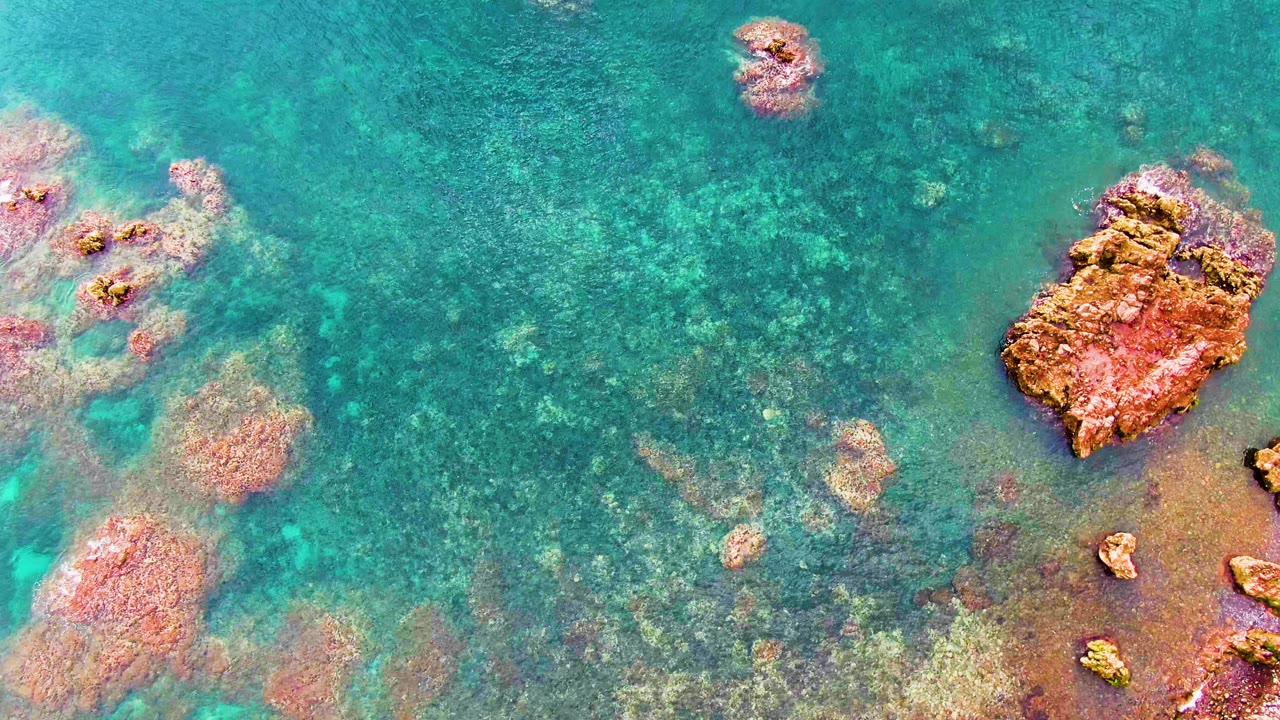 Overhead Santa Rosalia coastal water view with a large rust-colored rock near the top right, smaller submerged rocks, and no visible people.