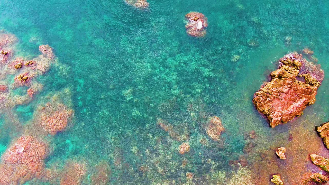 Overhead view of clear turquoise water in Santa Rosalia with a large reddish-brown rock near the right edge and patchy seabed color visible below, no visible people.
