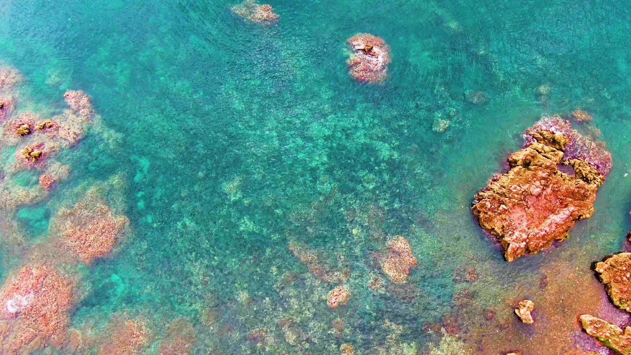 Overhead Santa Rosalia frame with scattered submerged rocks, pale seabed patches, and a larger reddish rock entering the lower right, no visible people.