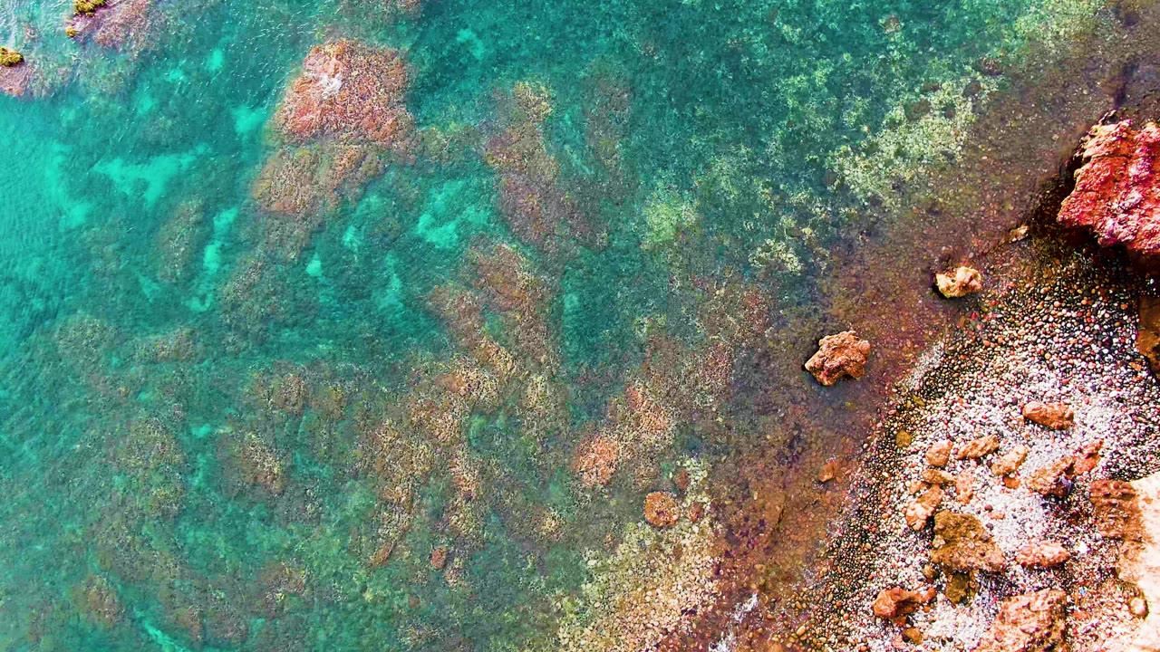 An overhead frame shows turquoise shallows, darker submerged rock patches, and reddish coastal rocks along the right edge in Santa Rosalia, Mexico, with no visible people.