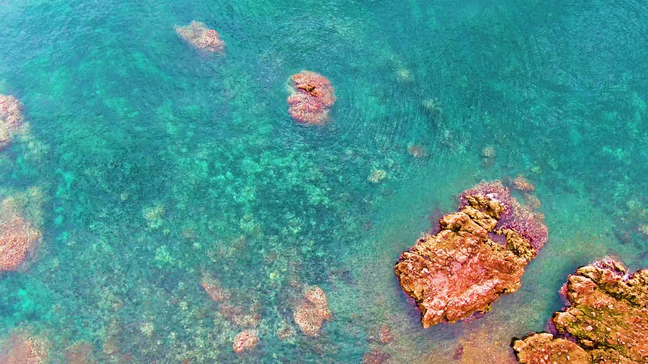 Overhead view in Santa Rosalia, Baja California Sur, showing clear turquoise water, mottled submerged rocks, reddish-brown rocks along the lower edge, and no visible people.