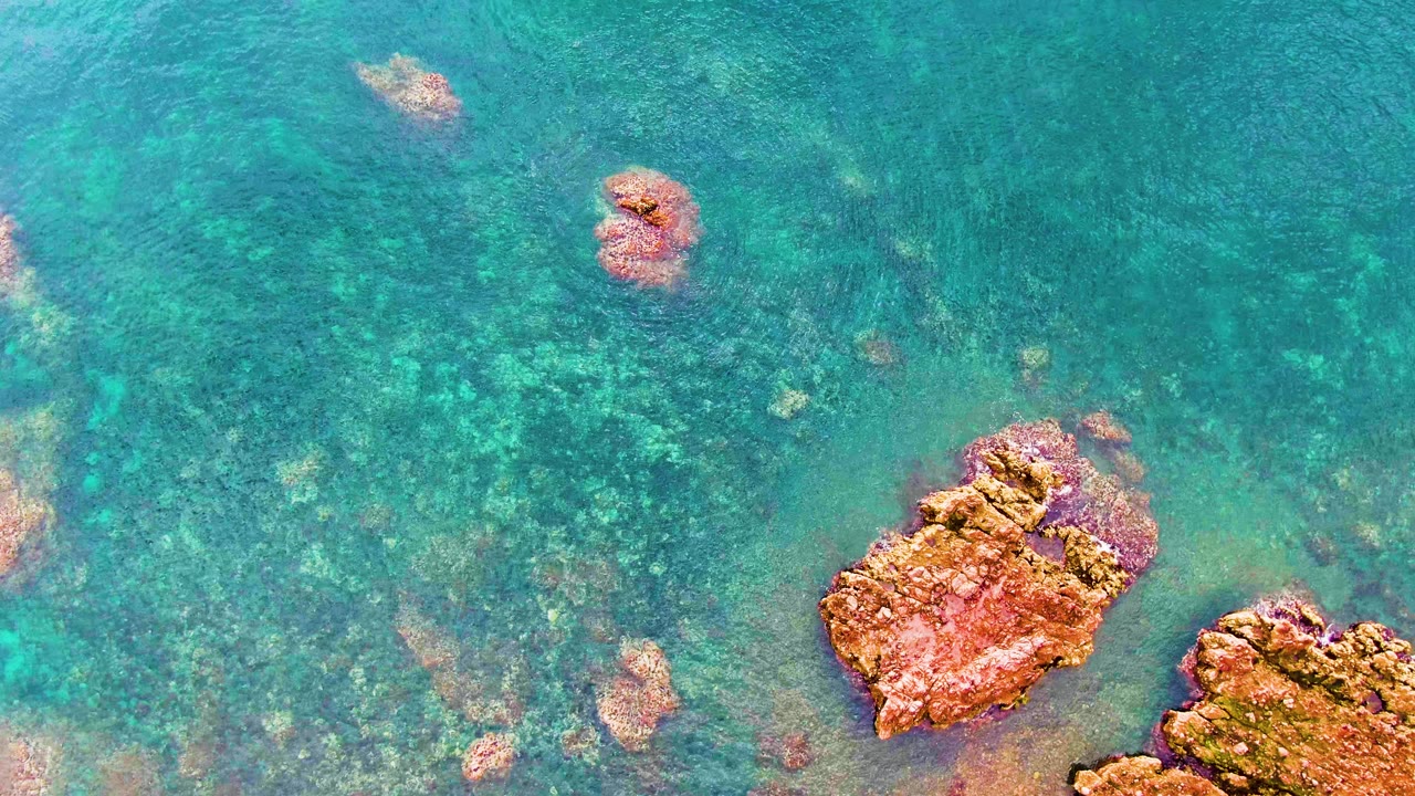 Second frame from Santa Rosalia shows blue-green shallow water with a broad patch of rocky seabed, two reddish rock masses near the bottom edge, and no visible people.