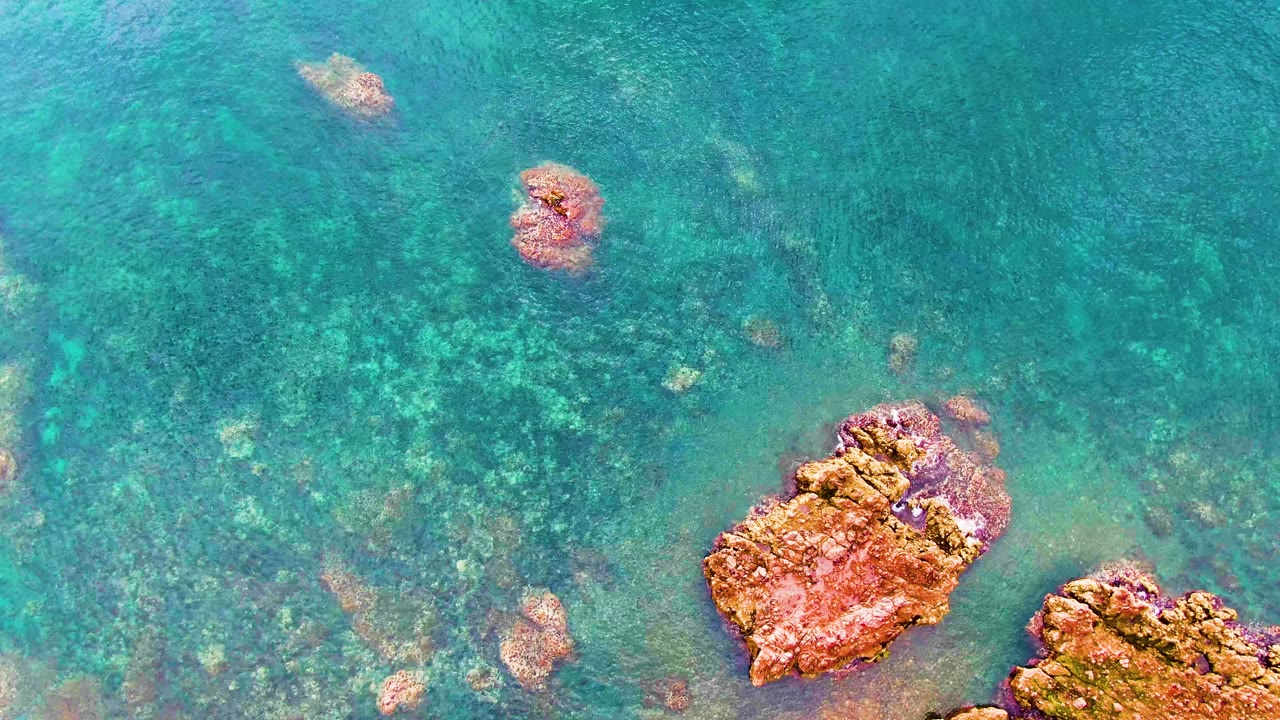 Overhead Santa Rosalia frame with turquoise water filling the view, scattered darker underwater rock shapes, reddish rocks at the lower right, and no visible people.