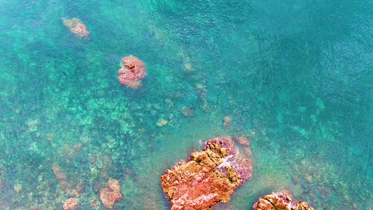 Top-down view of clear turquoise water in Santa Rosalia, Baja California Sur, with orange rocks near the lower edge, smaller submerged rocks above, and no visible people.