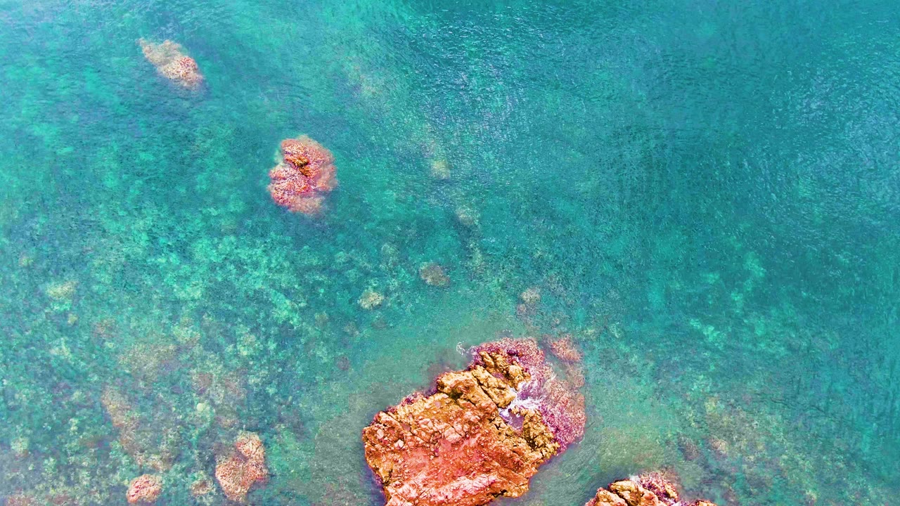 Clear blue-green ocean in Santa Rosalia with a large orange rock closer to the bottom center, scattered submerged rocks, shifting ripple patterns, and no visible people.