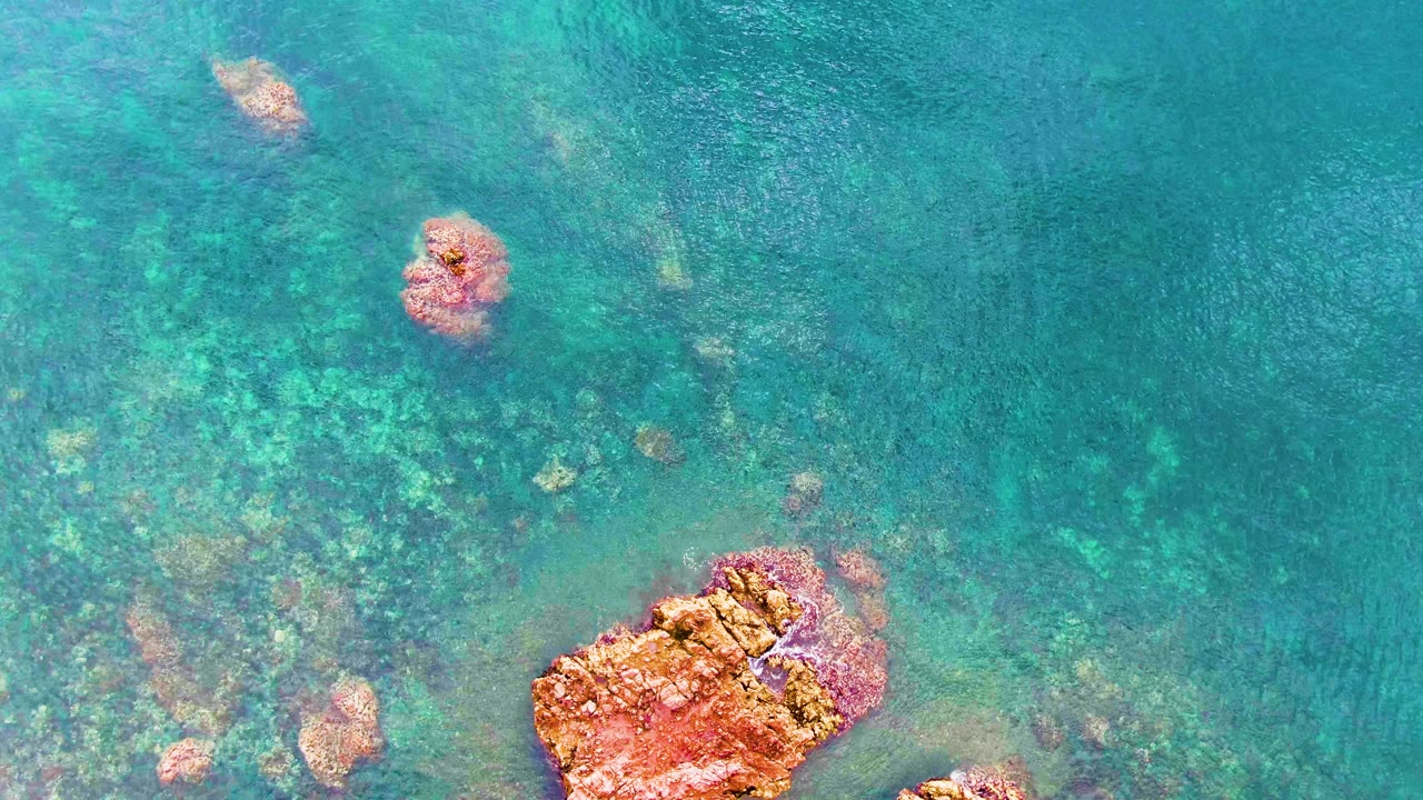 Aerial view over Santa Rosalia turquoise water, with the largest orange rock near the lower right, faint green seabed patches, small rock heads, and no visible people.