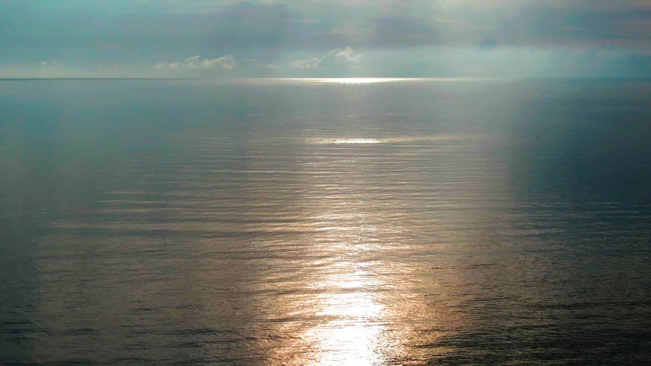 A brighter band of reflected sunlight spreads down the blue-gray water near Santa Rosalia, Mexico, with clouds above and no visible people.