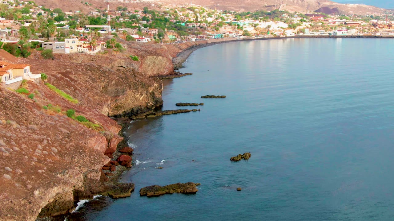 Aerial view of Santa Rosalia, Baja California Sur, with red-brown cliffs on the left, calm blue water on the right, dark offshore rocks, cliff-top buildings, and no visible people.