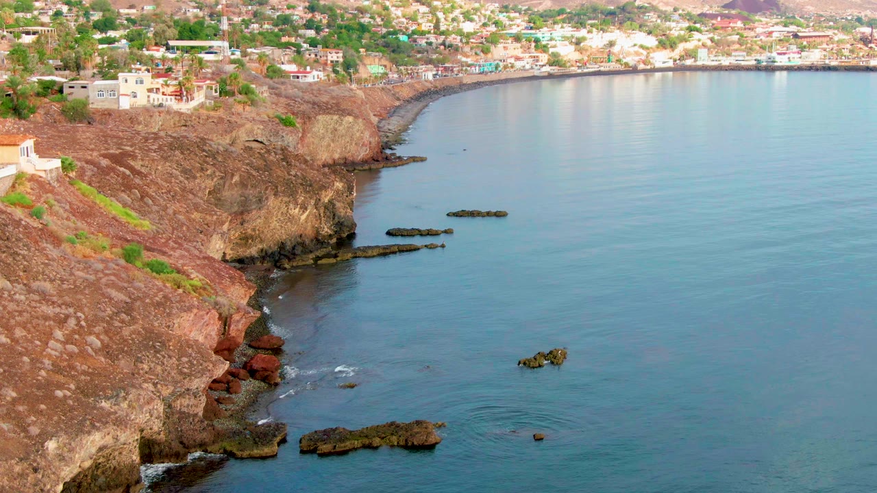Santa Rosalia coastline from above, showing the cliff face closer to center, small white wave breaks near the rocks, buildings along the upper slope, and no visible people.