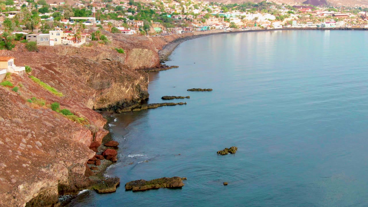 Rugged cliff edge with dark rocks and gentle surf below town buildings.