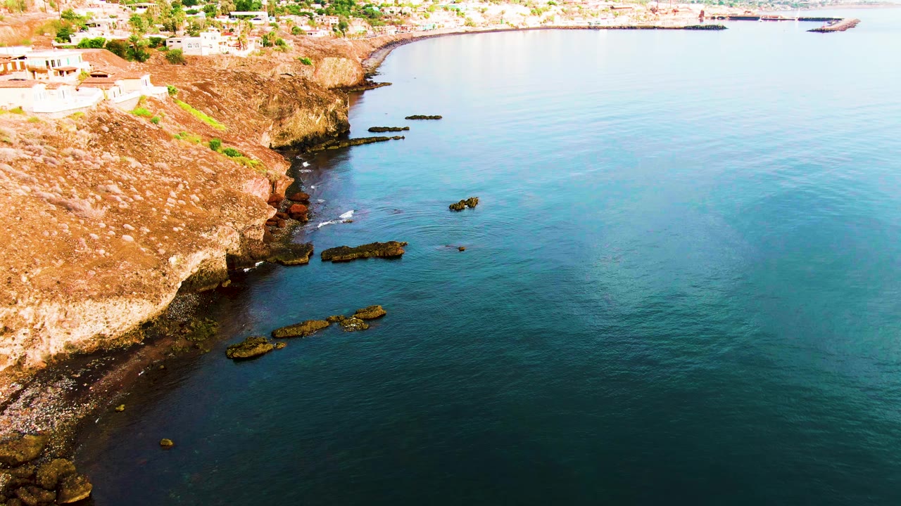 Opening aerial frame of Santa Rosalia coastline with brown rocky cliff on the left, calm blue-green ocean to the right, small buildings above the shore, and no visible people.