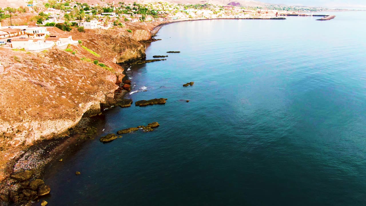 Aerial frame in Santa Rosalia showing the rocky bluff along the left side, glassy blue water across the right, cliff-top buildings, two distant boats, and no visible people.