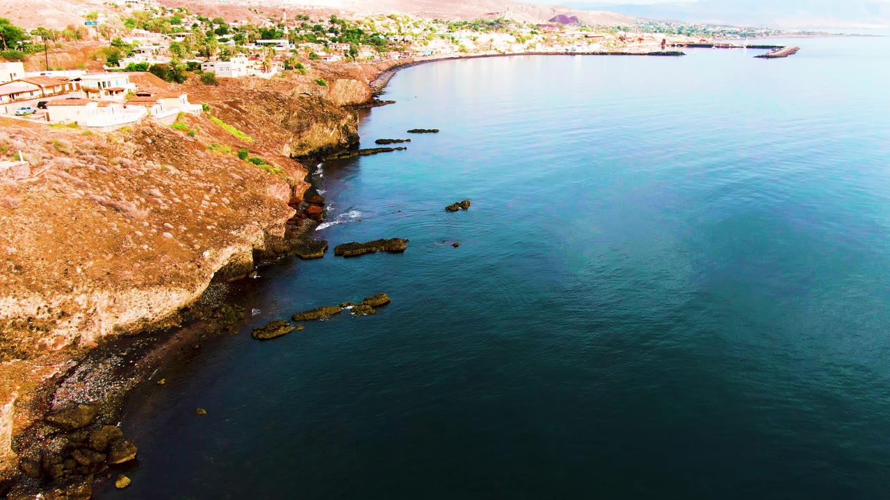 Aerial view over Santa Rosalia's rocky coast with brown cliffs, dark rock shelves in the water, pale buildings above the shoreline, distant boats, and no visible people.