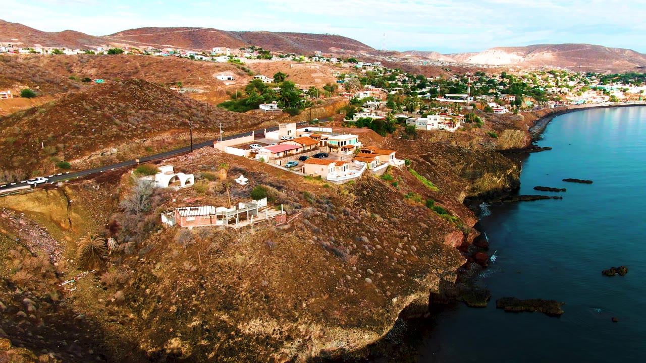 Aerial frame over Santa Rosalia, Baja California Sur showing brown hillside, residential buildings, a road, blue shoreline water, distant mountains, and no visible people.
