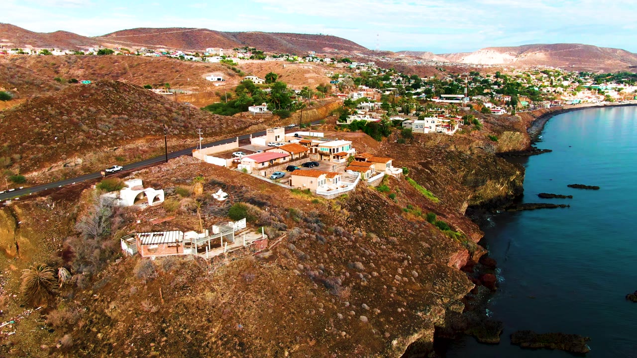 Santa Rosalia aerial frame with the hillside buildings slightly farther along the coast, parked cars near the structures, the road at left, water at right, and no visible people.