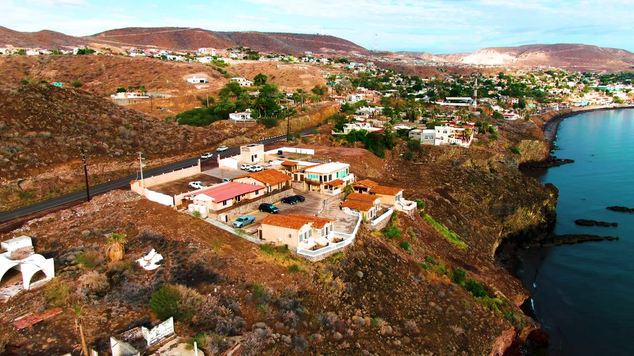 Opening aerial frame over Santa Rosalia, Baja California Sur, with red-roofed buildings on a dry hillside, a road above them, ocean to the right, and no visible people.